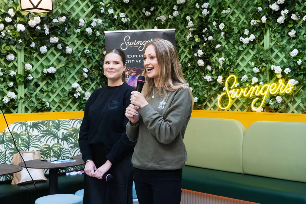 Two women speaking in front of a flower wall and neon Swingers sign